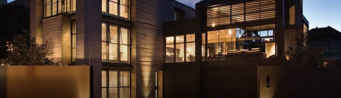 Modern house illuminated at twilight with warm interior lights and exterior uplighting.
